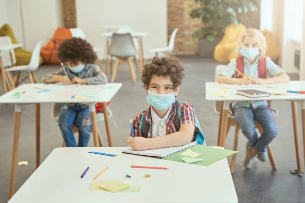 istock Happy school boy wearing face mask during coronavirus outbreak smiling while sitting at the desk in classroom. Kids studying in elementary school 1354694560