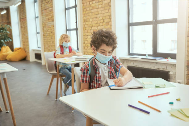 istock School boy wearing face mask during corona virus and flu outbreak writing in his notebook while sitting at the desk in a classroom. Kids studying in elementary school 1354694562