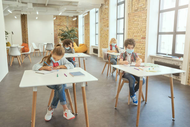 istock Studying during pandemic. School girl wearing face mask writing in her notebook while sitting at the desk in a classroom. Group of kids in elementary school 1354694570