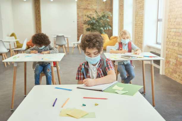 istock School boy wearing face mask during coronavirus outbreak writing in his notebook while sitting at the desk in classroom. Kids studying in elementary school 1354694572