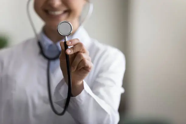 Photo of Happy young GP doctor using stethoscope, showing tool at camera