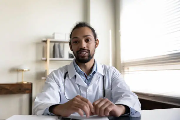 Empathetic young mixed raced doctor giving online help, medical support, therapy advice, consultation. Head shot portrait of male general practitioner looking, speaking at camera. Video call screen