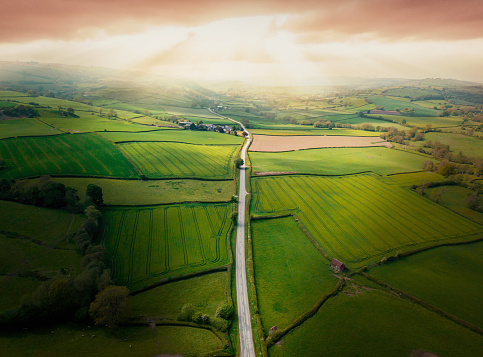 Aerial view looking down on a rural road in the UK countryside with a car racing along it. On a bright sunny day, farmland and crops can be seen either side of the road