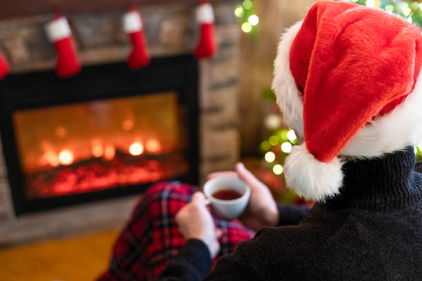 homme avec le café de tasse, le thé dans le chapeau de père noël se reposant et se chauffant au soir d’hiver près de la flamme de cheminée, plaid couvert de noël - holiday emotional stress christmas santa claus photos et images de collection