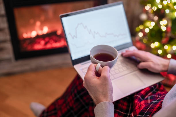 istock Man freelancer working on laptop with graphs and charts on screen sitting near christmas tree and fireplace with cup of tea. 1354711178