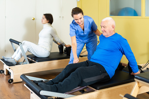 Senior man practicing pilates system on reformer in rehabilitation center to improve and maintain mobility under supervision of qualified female doctor