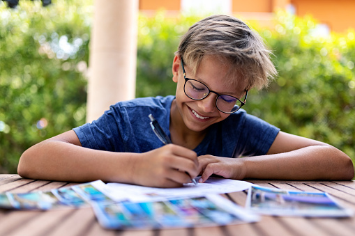 Teenager boy writing postcards to friends and family. He is sitting by a table in the backyard. Toothy smile, one person, laughing\nCanon R5