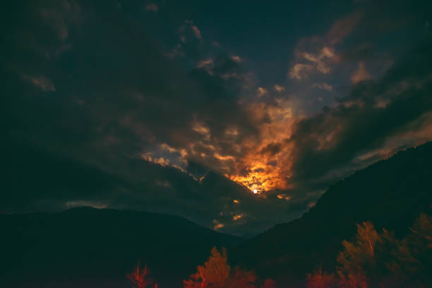 la luna piena arancione che sorge da dietro la catena montuosa illumina le nuvole nebbiose, creando un'atmosfera mistica.superluna.la luna nascente nel cielo notturno con strati di colline in primo piano - crateri foto e immagini stock