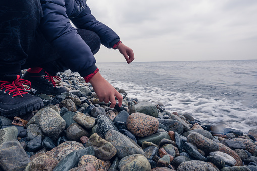 Unrecognizable boy looking for a stone for stone skipping on a stony beach on an cloudy autumn day. Close-up with copy space.
