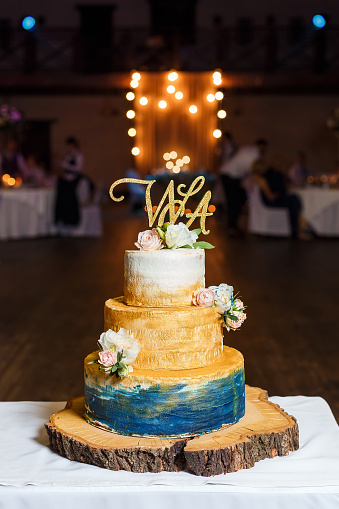 Elegant wedding cake in gold and blue colors decorated with fresh flowers and gold topper, standing on big piece of tree cut.