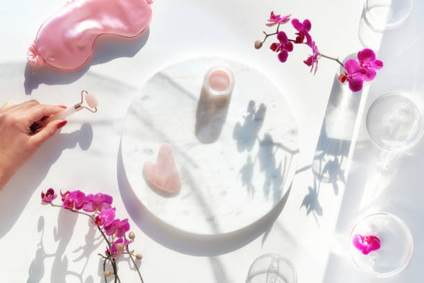 istock Pink quartz face roller in hand, moisturizing cream on marble plate. Exotic flat lay with monstera leaves and vibrant magenta orchid flowers. Sunlight, long shadows. Facial massage 1354729623