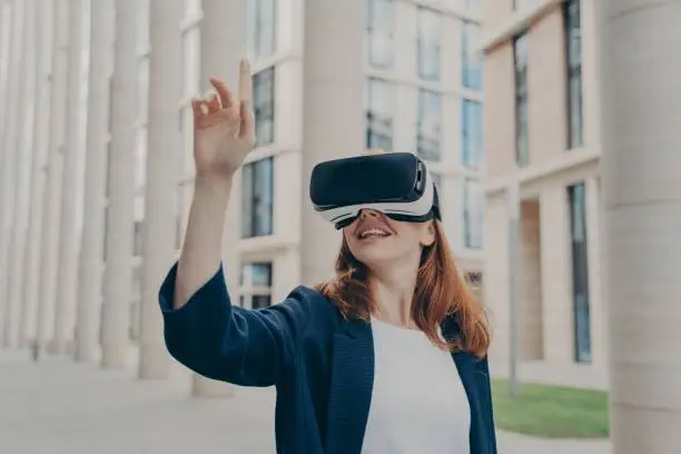 Business meeting in VR. Cheerful redhead female dressed formally stands outdoors in virtual reality goggles and interacts with virtual interface, pressing invisible buttons, moving forefinger in air