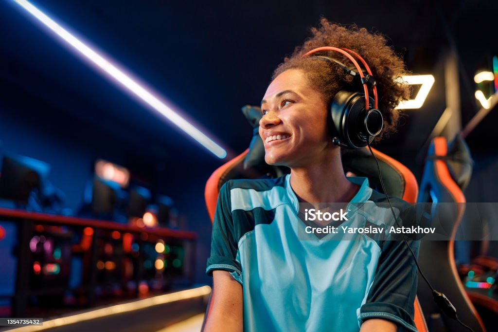 Female gamer relaxes after tournament in cyber club African female wearing wired headphones looking to side while sitting on gaming chair with happy facial expression in computer club Gamer Stock Photo