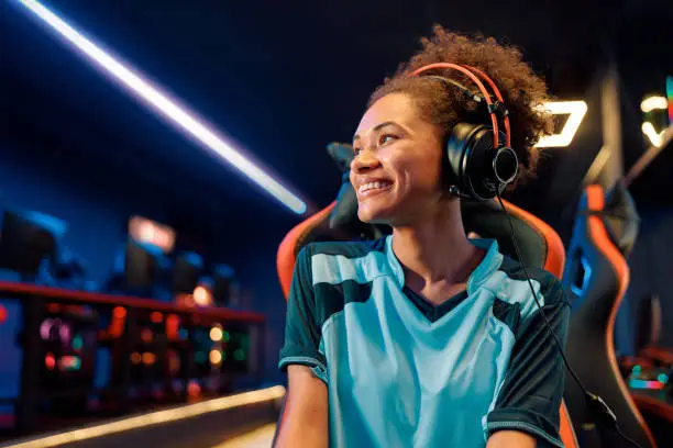 African female wearing wired headphones looking to side while sitting on gaming chair with happy facial expression in computer club