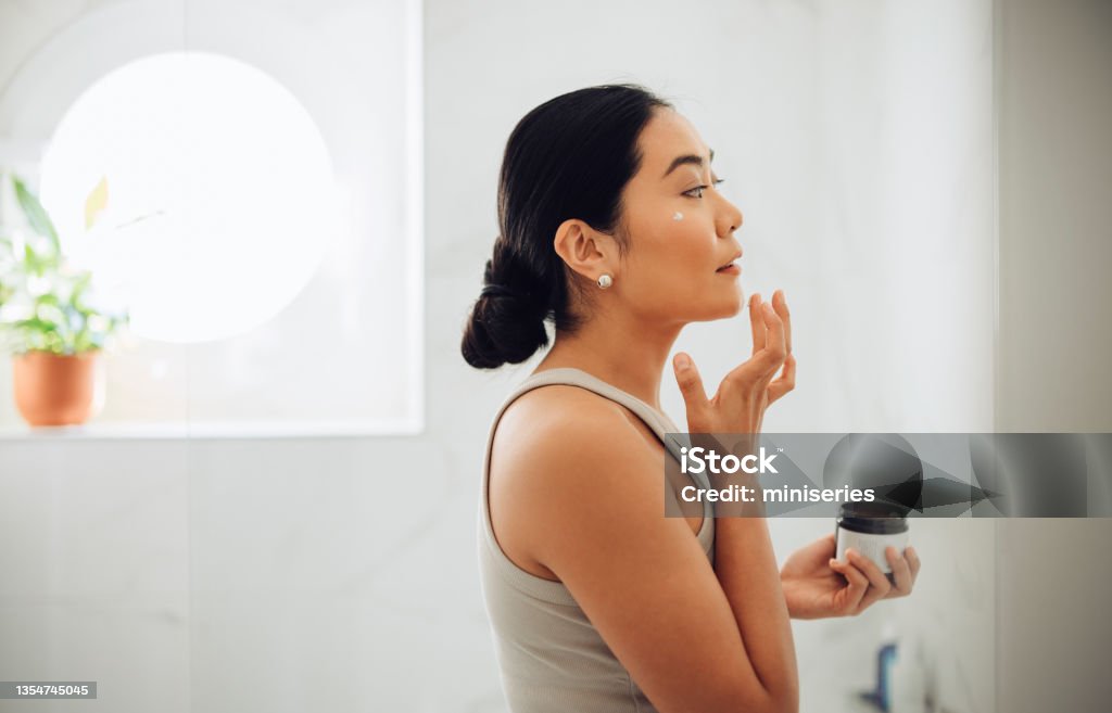 Morning Routine: Attractive Asian Woman Applying Face Cream in her Home Beautiful and cheerful Asian woman standing in her bathroom, holding face cream and applying it to her cheeks and face. Women Stock Photo