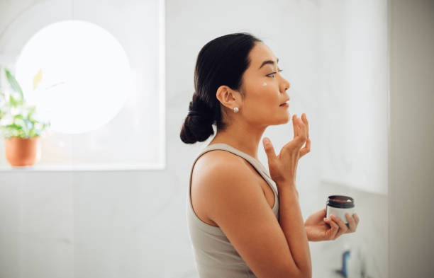 Morning Routine: Attractive Asian Woman Applying Face Cream in her Home Beautiful and cheerful Asian woman standing in her bathroom, holding face cream and applying it to her cheeks and face. applying stock pictures, royalty-free photos & images