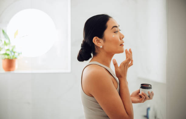 Morning Routine: Attractive Asian Woman Applying Face Cream in her Home Beautiful and cheerful Asian woman standing in her bathroom, holding face cream and applying it to her cheeks and face. makeup product photography stock pictures, royalty-free photos & images