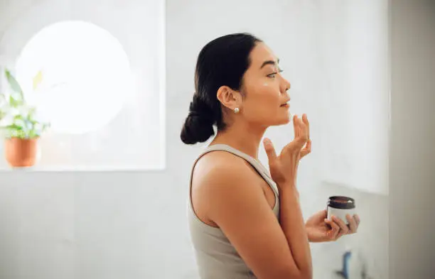 Beautiful and cheerful Asian woman standing in her bathroom, holding face cream and applying it to her cheeks and face.