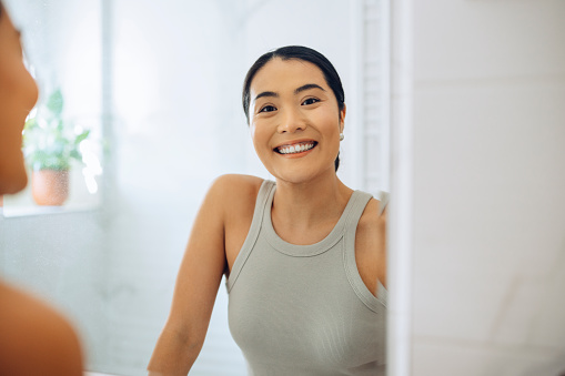 Cheerful Asian Woman standing at her sink in the bathroom, looking at herself in the mirror, smiling and holding a beauty care product.