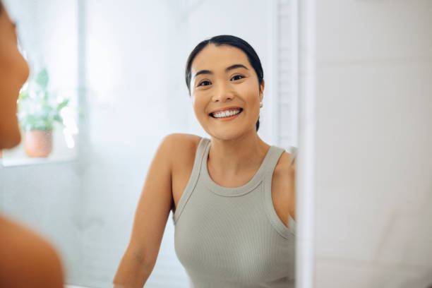 Morning Routine: Portrait of a Beautiful Asian Woman Looking at herself in the Mirror Holding a Beauty Care Product Cheerful Asian Woman standing at her sink in the bathroom, looking at herself in the mirror, smiling and holding a beauty care product. central asian ethnicity photos stock pictures, royalty-free photos & images