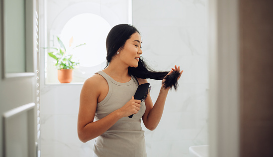 Beautiful Asian woman standing in her bathroom holding a hairbrush, brushing her hair.