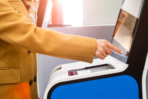 Woman passenger uses self service kiosk with touchscreen to check-in for flight to avoid long queue in contemporary airport terminal close side view