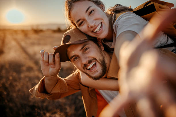 Selfie Portrait of cheerful couple having fun while piggybacking on field during sunset 20 29 years stock pictures, royalty-free photos & images