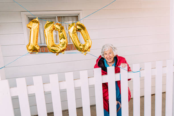 陽気な高齢の白人女性がフェンスに誇らしげに傾き、金色の風船で彼女の100歳の誕生日を祝う広角ショット - 114 ストックフォトと画像