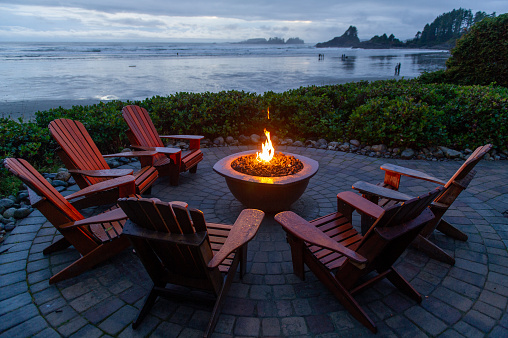 Adirondack chairs around a fire pit facing the beach of Cox Bay in Tofino Vancouver Island Canada. People are walking on the beach