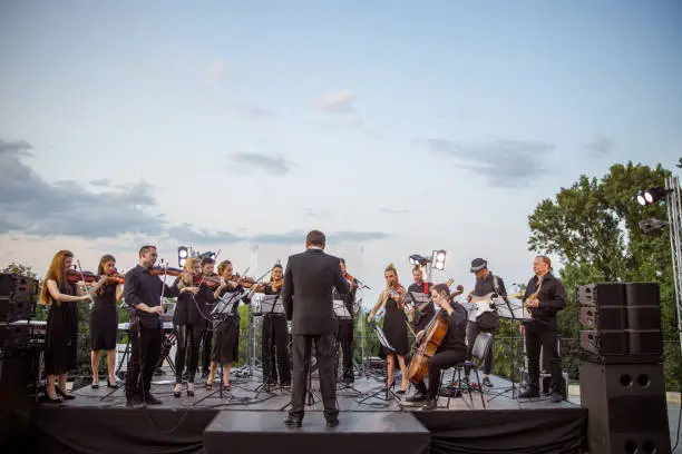 Musicians standing on outdoor stage and playing musical instruments while conductor leading orchestra performance under blue cloudy sky