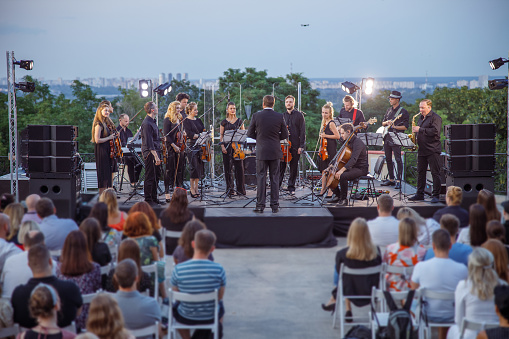 Conductor directing orchestra performance while musicians playing on outdoor stage and audience sitting on concert chairs