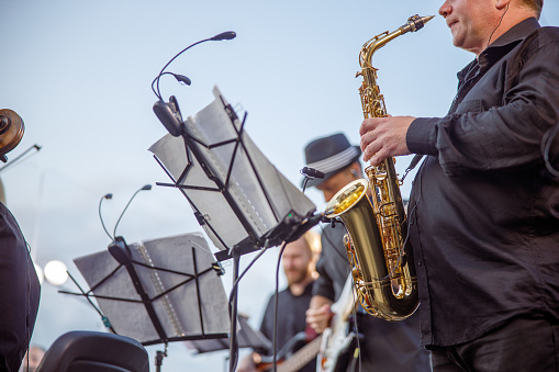 Man saxophonist standing near music stand with notes and playing saxophone while having concert rehearsal with musicians under blue sky