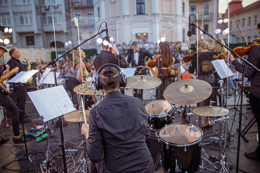 Back view of man drummer in headphone sitting at drum kit performing concert with musical ensemble on the street