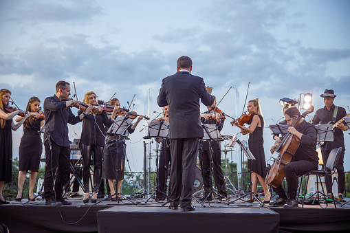 Musicians standing on outdoor stage and playing musical instruments while conductor leading orchestra performance