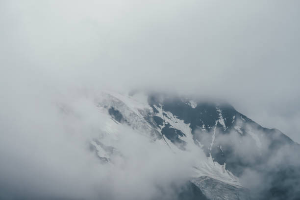 istock Minimalist monochrome atmospheric mountains landscape with big snowy mountain range in low clouds. Awesome minimal scenery with glacier on rocks. Black white high mountain ridge with snow in clouds. 1354775007