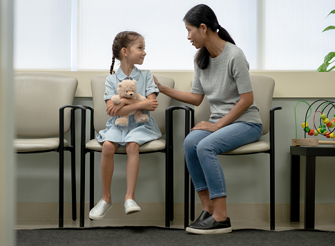 A little girl sits with her mother in the waiting room of a doctors office, as she clings to her teddy bear.  She is dressed casually in a light blue dress as her mother places her hand on the little girls shoulder and tries to reassure her that it will be okay.