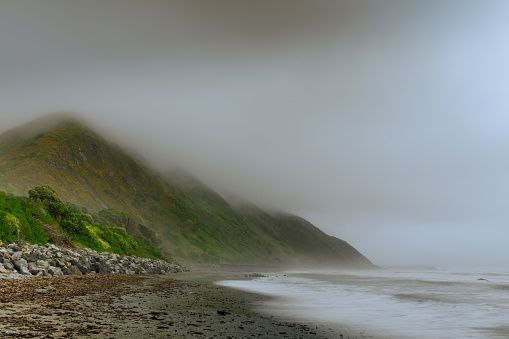 This October 2021 composite, long-exposure image shows the Tasman Sea washing up at Paekākāriki, along the Kāpiti Coast in Aotearoa New Zealand. The tops of the mountains are obscured by storm clouds. A rocky seawall is on the left. While the community's seawall has failed in recent storm surges, this image is not suitable for editorial use as buildings and a railroad line have been digitally removed. A seperate image is available.