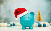 Teal piggy bank wearing Santa hat with Christmas decorations background. Christmas savings or spending theme