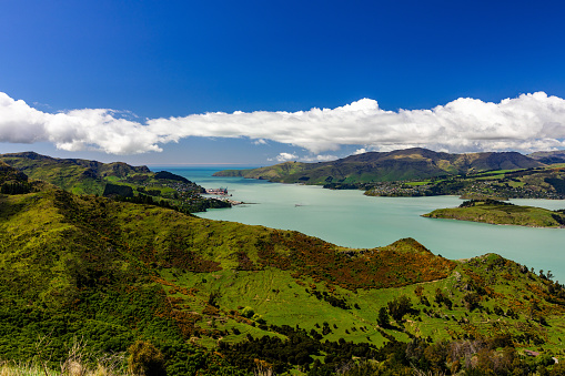 This October 2021 daylight image shows Te Whakaraupō Lyttelton Harbour and its teal waters. The cargo cranes at the port of Lyttelton are visible. The community of Diamond Harbour is on the opposite side of the harbour from the port. Te Moana-nui-a-Kiwa Pacific Ocean appears in the distance.  This area is part of Horomaka Banks Peninsula in Ōtautahi Christchurch, Aotearoa New Zealand. Most marine traffic has been digitally removed.