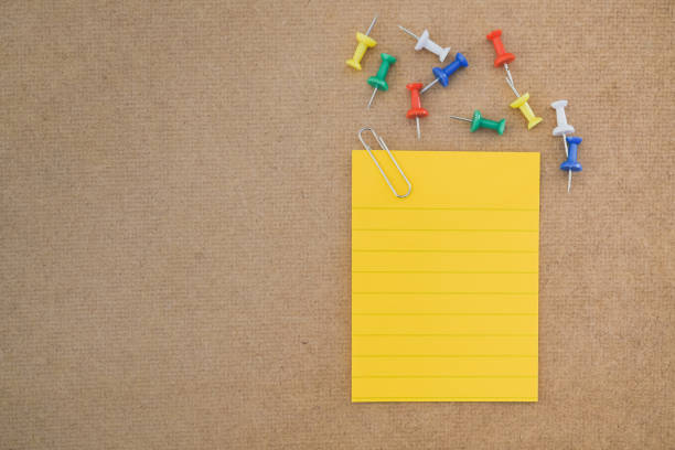 yellow note paper with paper clip and multi color thumbtacks on brown grunge board, top view