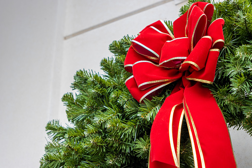 A traditional Christmas wreath with a giant red bow hangs on the outside of a white building door during the day.