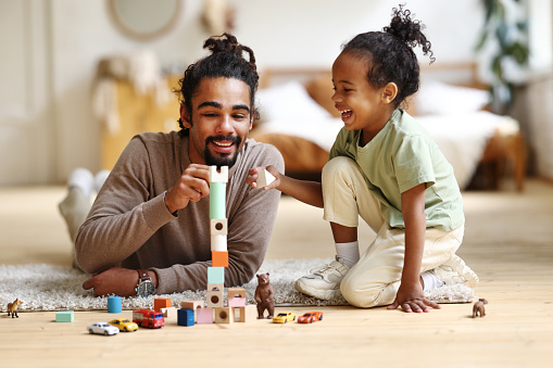 Happy african american family father and child son playing toys together at home, positive dad parent and little kid sitting on floor and stacking wooden pyramid, laughing while spending leisure time