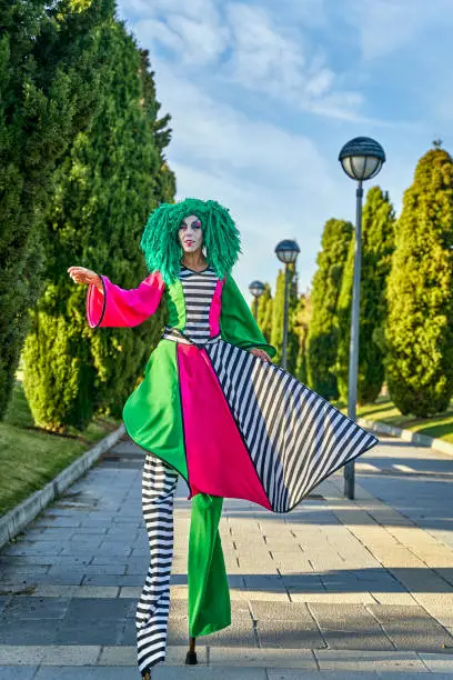 Full body of female stilt walker in funny harlequin costume and green wig strolling on alley amidst lush trees during parade in sunny park