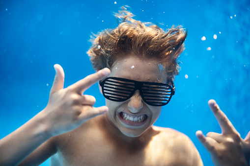 Teenage boy wearing shutter shades underwater. The boy is showing rock and roll gesture to the camera.\nCanon R5