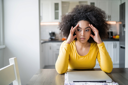 African american young woman dealing with financial problems and bills at home, sitting at the kitchen table, with closed laptop, feeling headache, stress and fatigue. Bad news, despair, overthinking