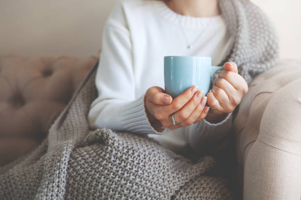 Close up of hands holding blue cup of tea or coffee. Model in white sweater and cozy plaid is sitting, relax at home on sofa. Depth of field and empty space on blurry beige background Close up of hands holding blue cup of tea or coffee. Model in white sweater and cozy plaid is sitting, relax at home on sofa. Depth of field, empty space on blurry beige background cup of tea hands stock pictures, royalty-free photos & images