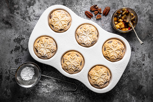 A white china muffin tray full of freshly baked mince pies. Each pie is decorated with an icicle and star made from pastry. They are dusted with icing sugar. A small sieve of icing sugar, a bowl of mincemeat and some pecan nuts sit beside them.