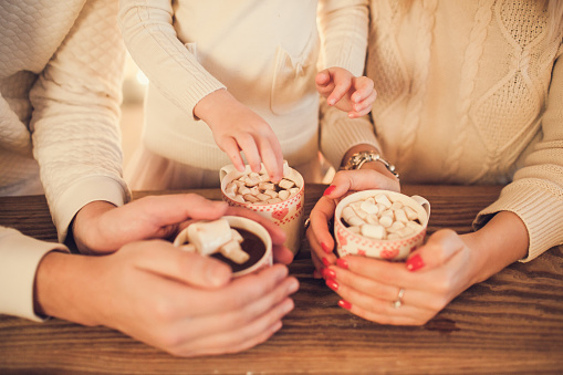 Family: mom, dad and daughter in white sweaters cook and drink cocoa with marshmallows. Closeup hands and cups. Christmas concept.
