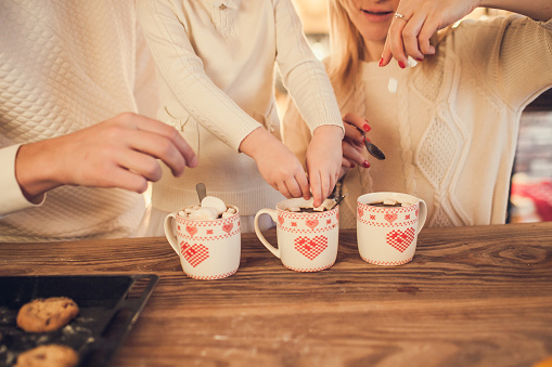 Family: mom, dad and daughter in white sweaters cook and drink cocoa with marshmallows. Closeup hands and cups. Christmas concept.