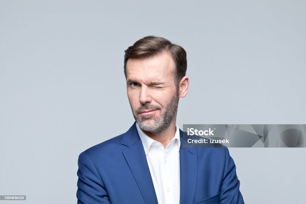 Headshot of cheerful mature businessman Portrait of mature man wearing navy blue jacket and white shirt, winking at camera. Studio shot of male entrepreneur against grey background. Winking Stock Photo